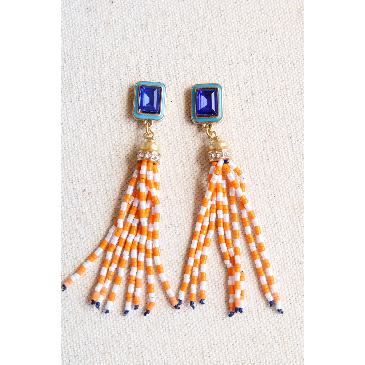 Pair of earrings with blue tops and orange and white striped fringes on a light background