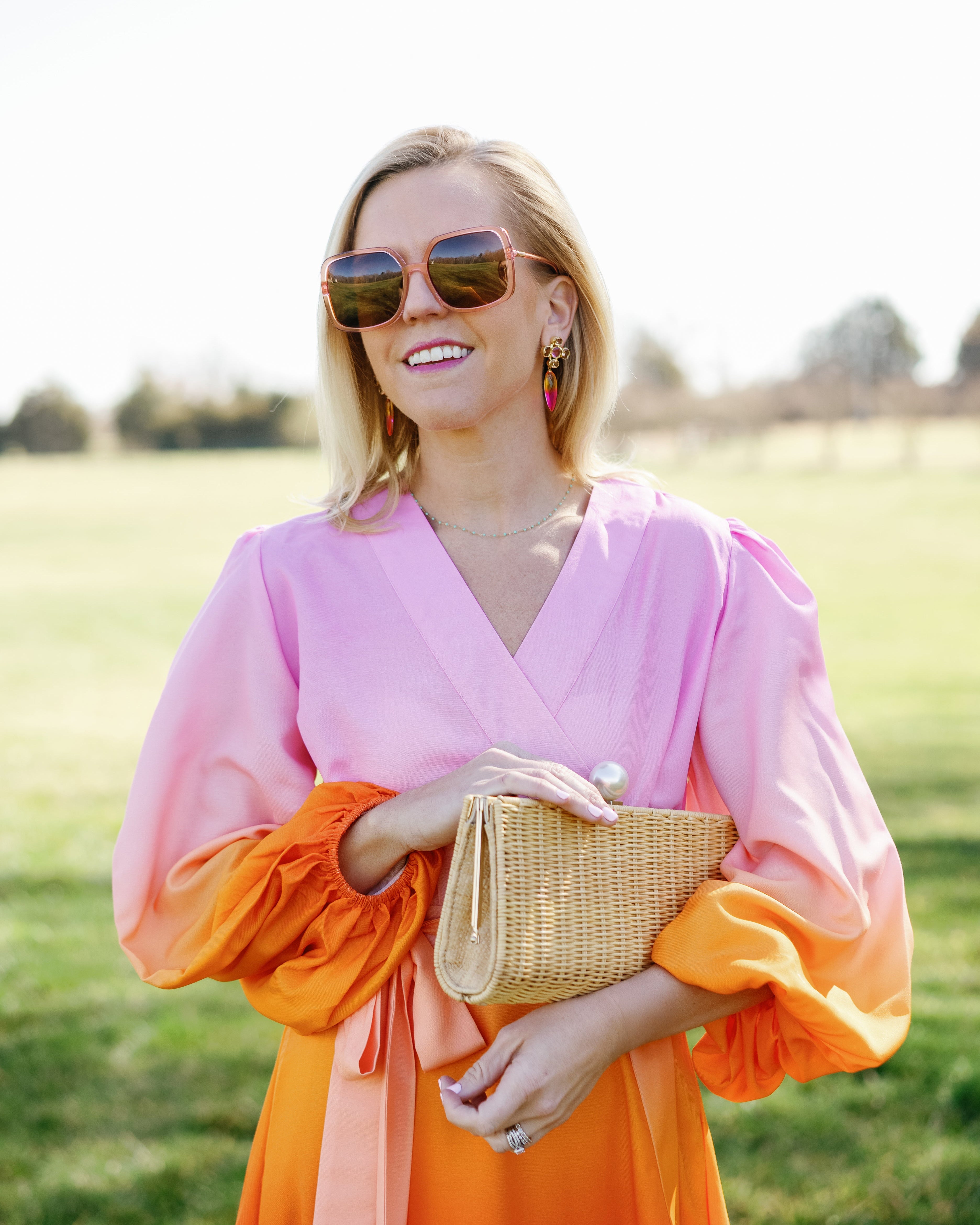 Woman in a pink and orange dress holding a gold clutch in a field