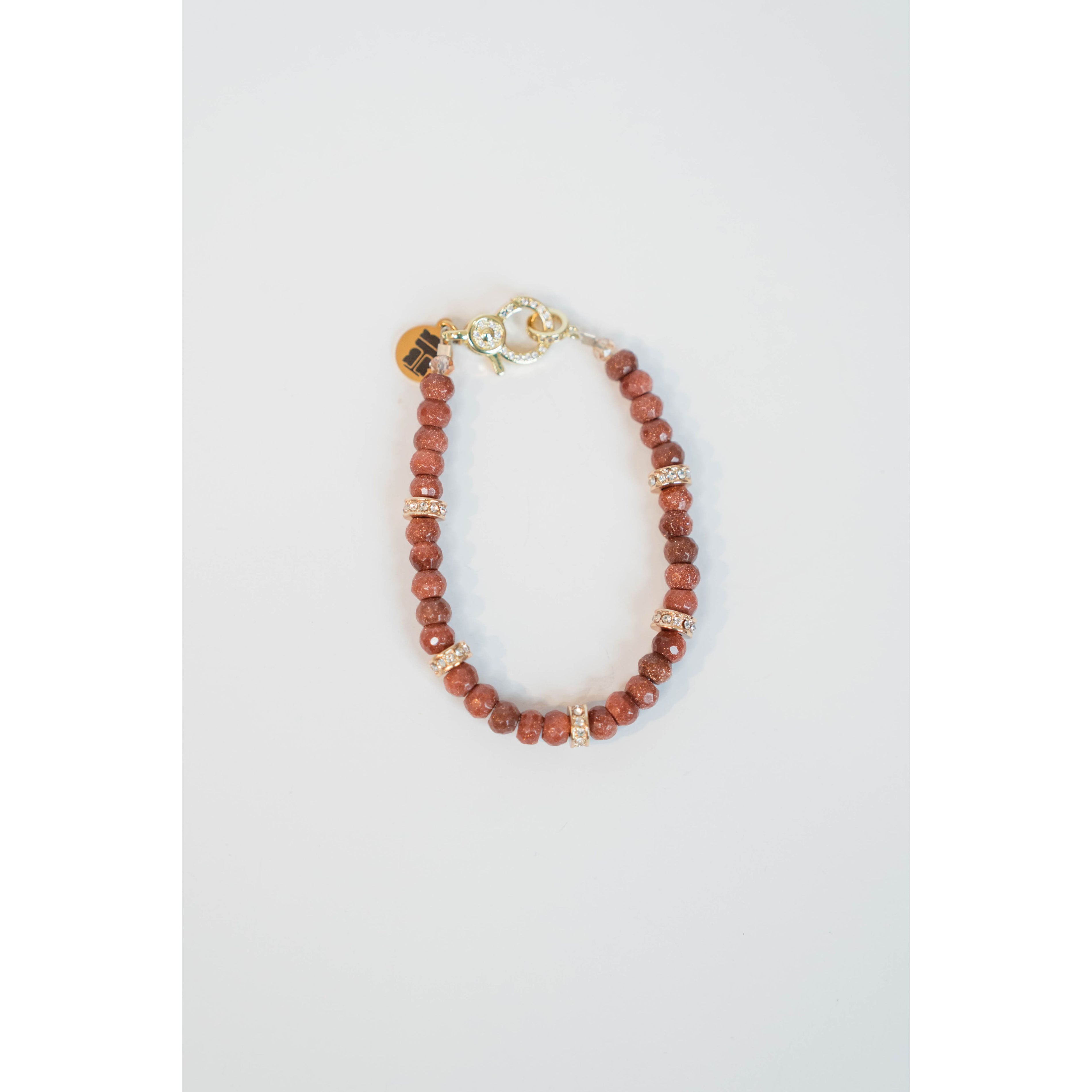 A bracelet with a pattern of goldstone and pavé beads are strung on cord with a pavé clasp and logo charm.