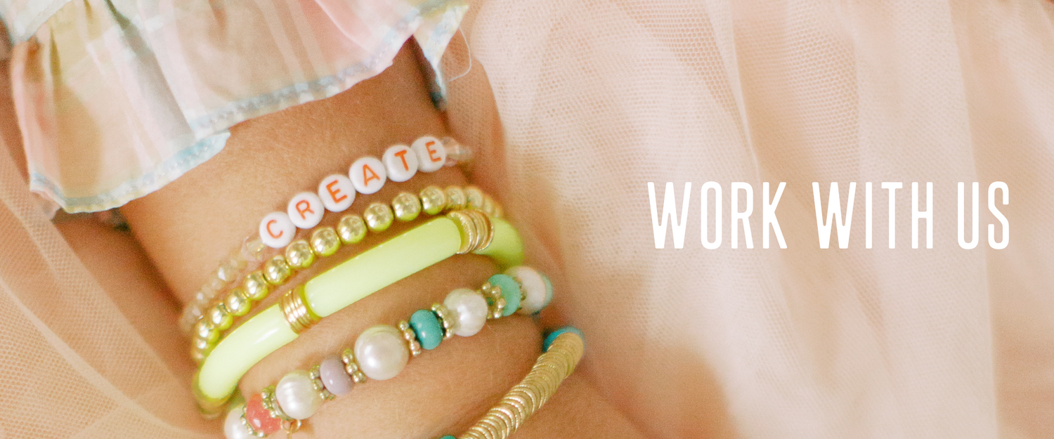 Close-up of colorful bracelets on a wrist, including one that spells 'CREATE,' with 'WORK WITH US' text on a soft pink background.