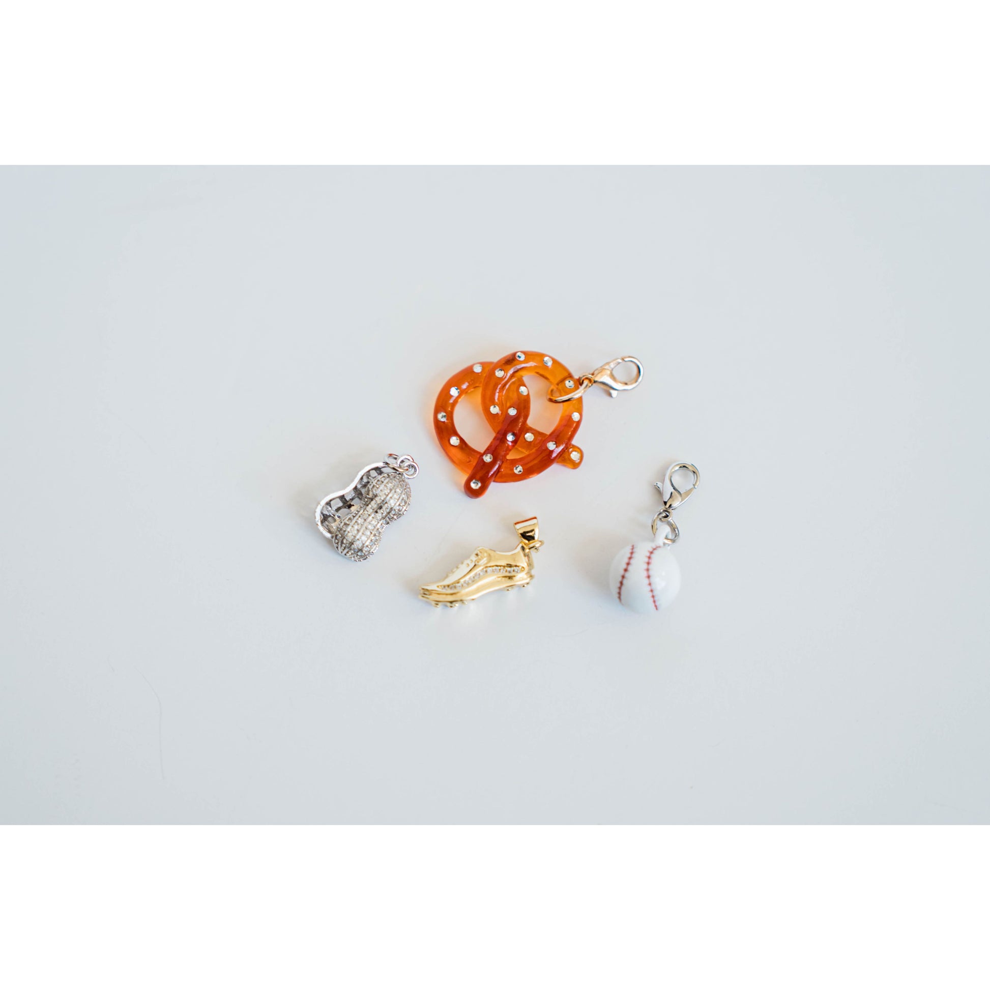 set of four baseball-themed charms featuring acrylic pretzel charm, silver pave peanut charm, golden baseball cleat with pave detailing, and white baseball charm