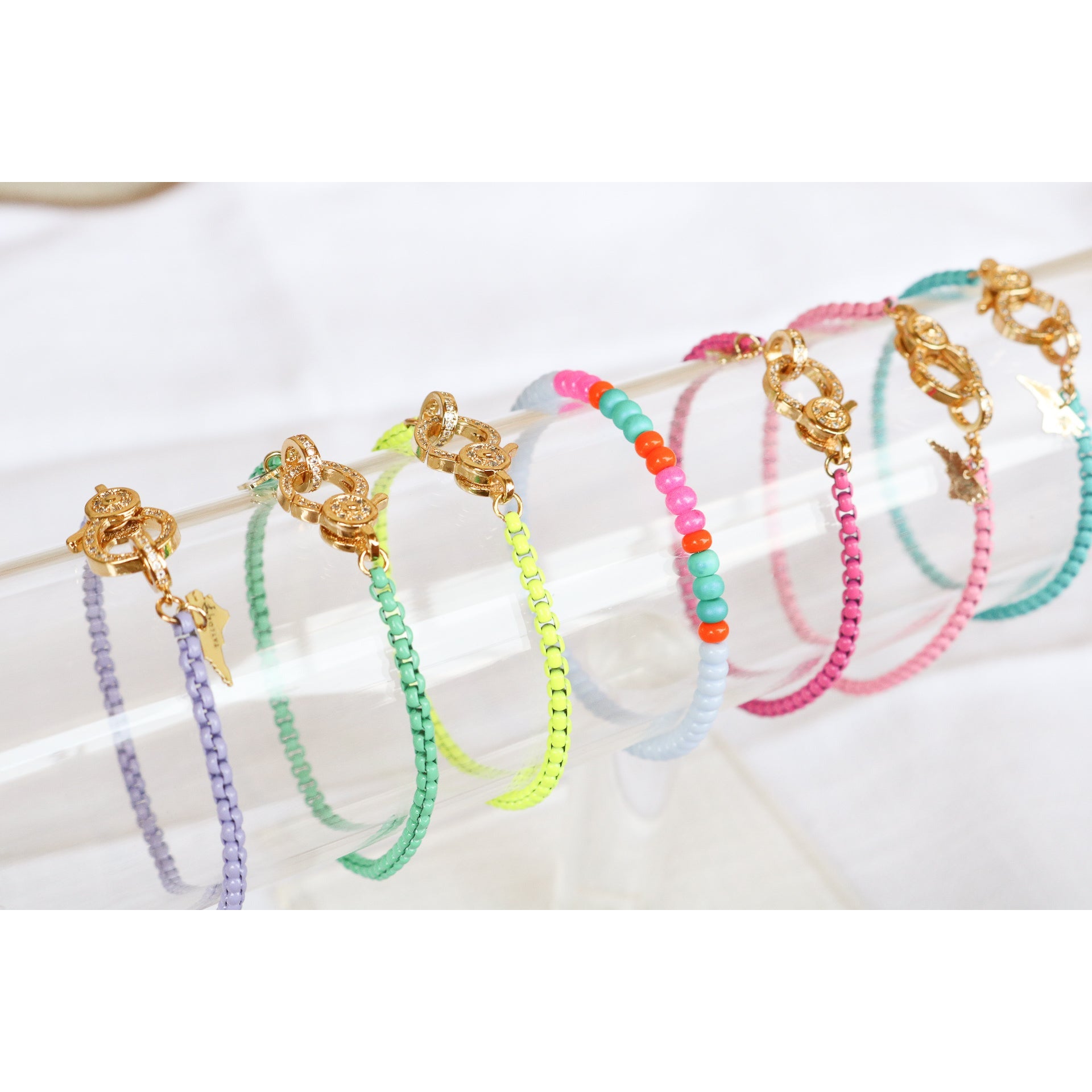 A collection of colorful enamel box chain bracelets with gold-tone clasp, displayed on a clear acrylic stand.