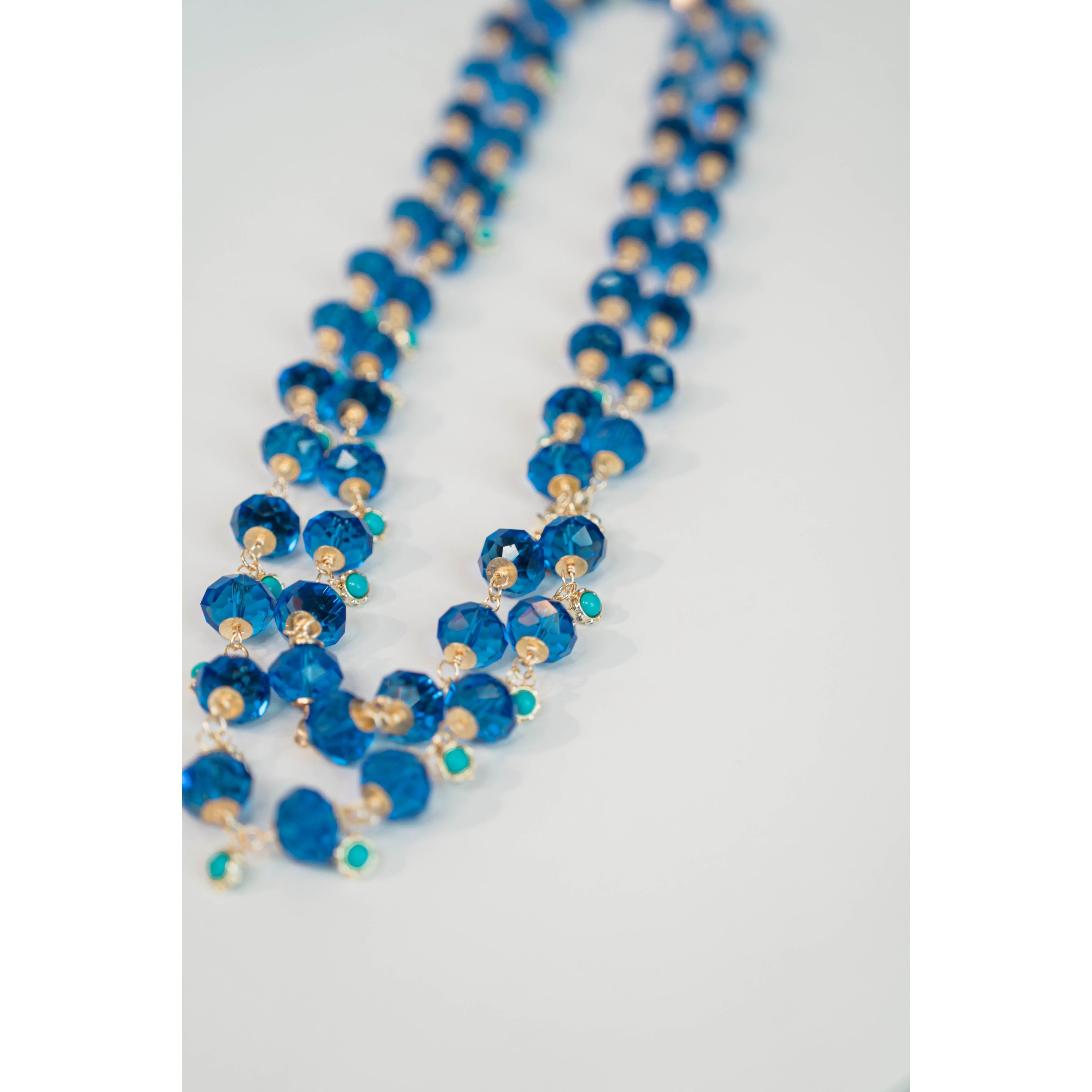 Long beaded necklace with crystal blue and turquoise and gold chain on a light gray background