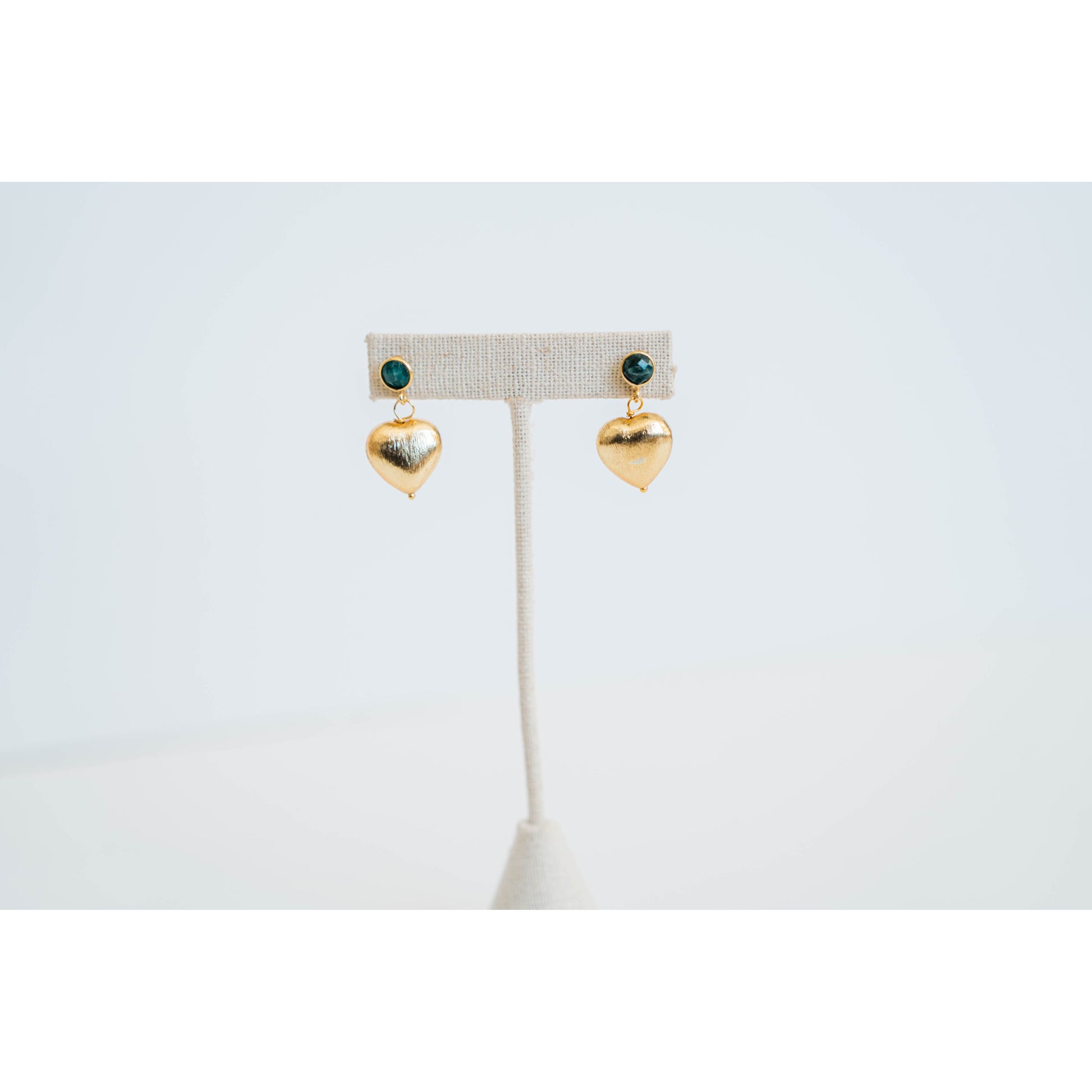Gold heart-shaped earrings with an emerald green stone stud on a white stand against a light gray background
