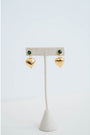 Gold heart-shaped earrings with an emerald green stone stud on a white stand against a light gray background