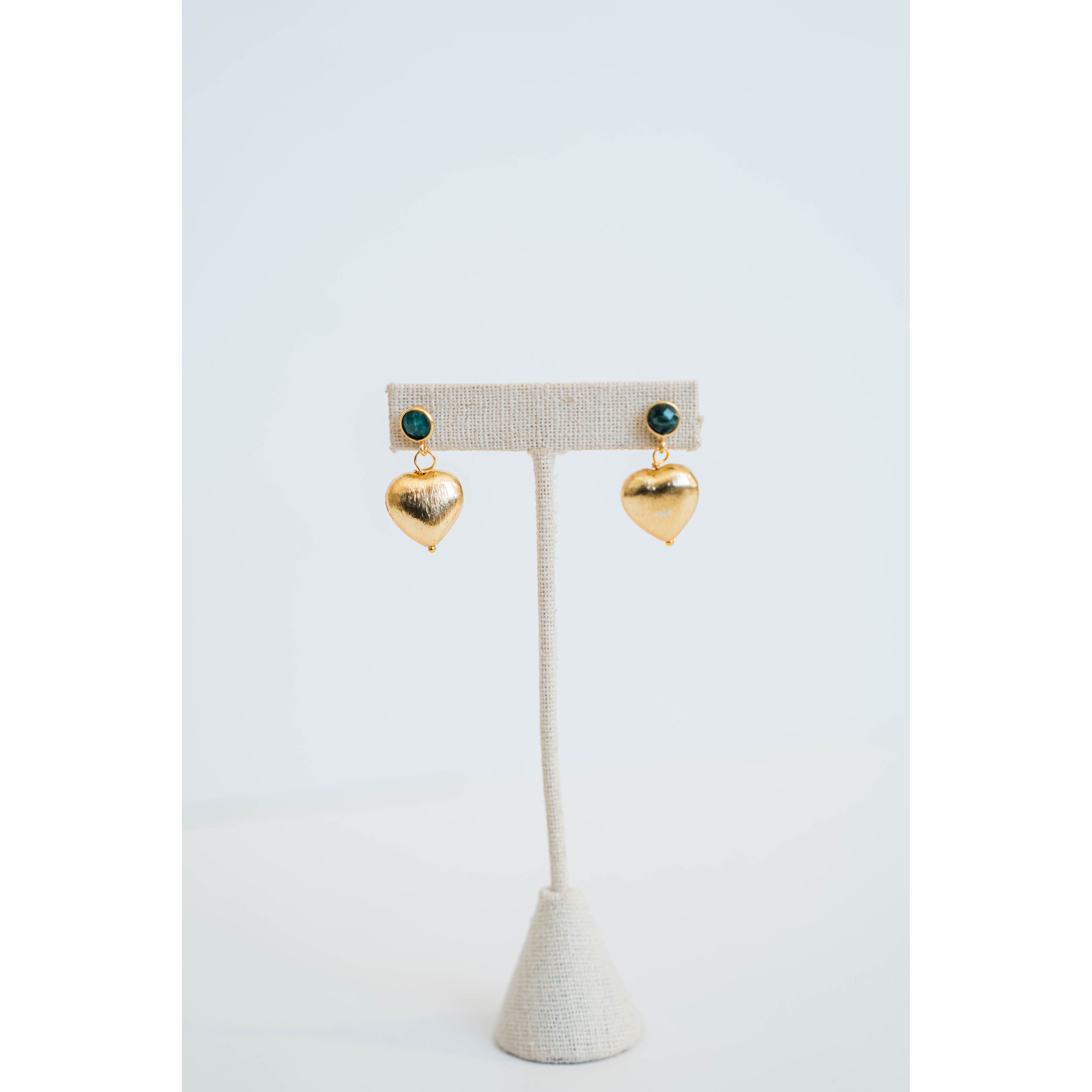 Gold heart-shaped earrings with an emerald green stone stud on a white stand against a light gray background