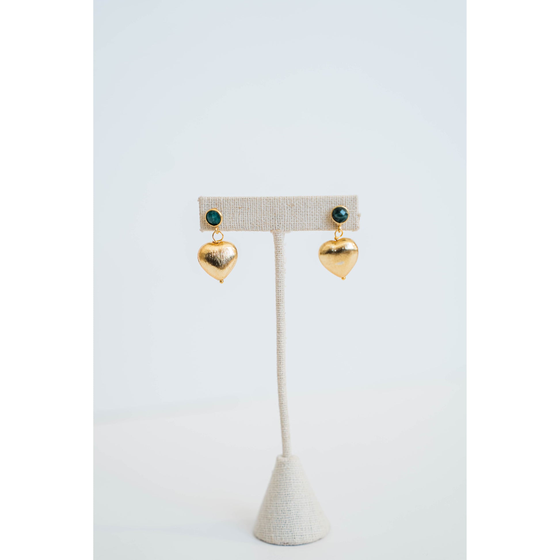 Gold heart-shaped earrings with an emerald green stone stud on a white stand against a light gray background