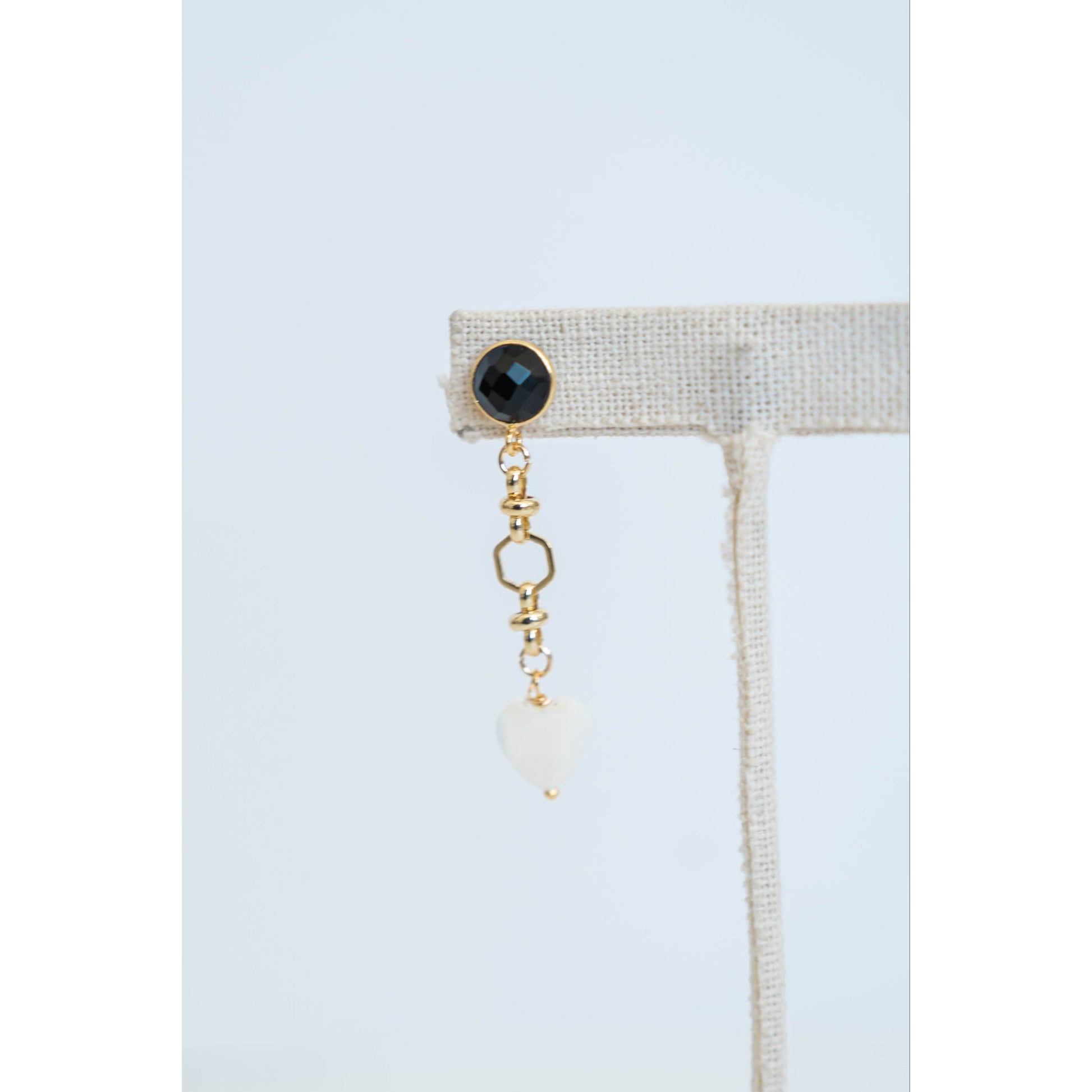 Gold chain earring with black gemstone and white heart pendant on a light gray background