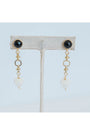 Pair of gold dangle earrings with black gemstones and white heart pendants on a light grey background