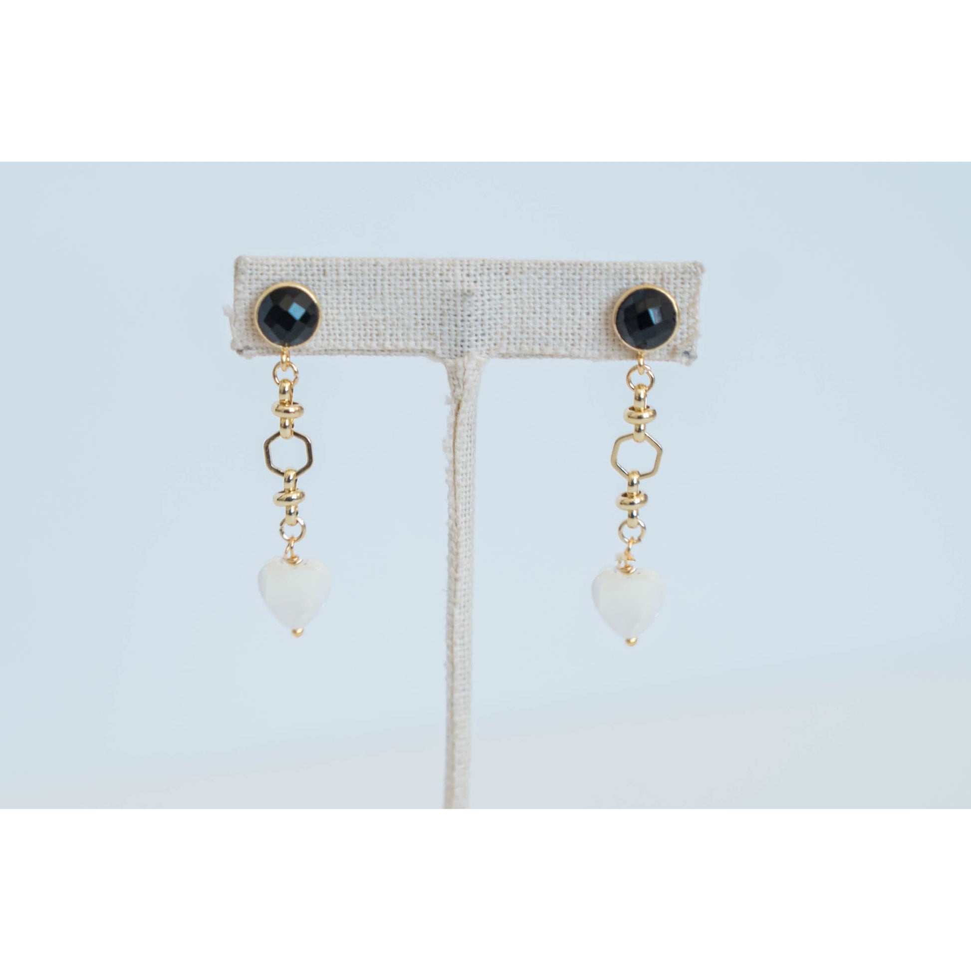 Pair of gold dangle earrings with black gemstones and white heart pendants on a light grey background