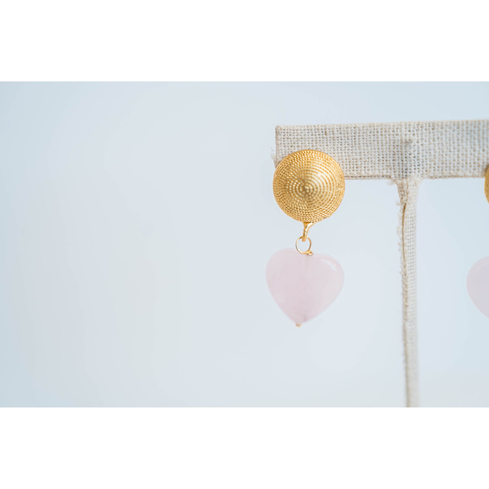 earring with large gold textured circular posts and  a light pink quartz heart dangle accent on an earring stand on a light background