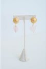 earrings with large gold textured circular posts and light pink quartz heart dangle accents on an earring stand on a light background