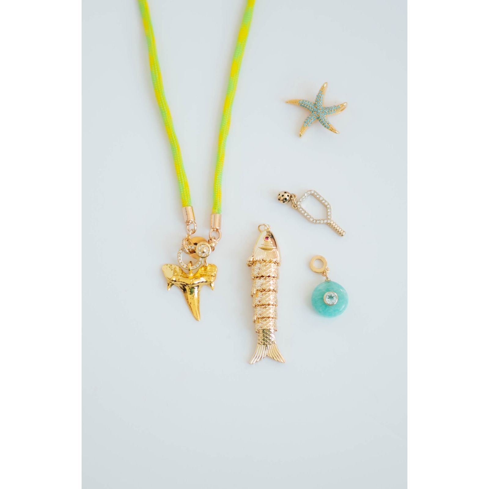 grouping of a gold fish pendant, shark tooth charm, starfish pendant, pave pickleball charm, and round turquoise charm with a neon yellow cord necklace against a light gray background.