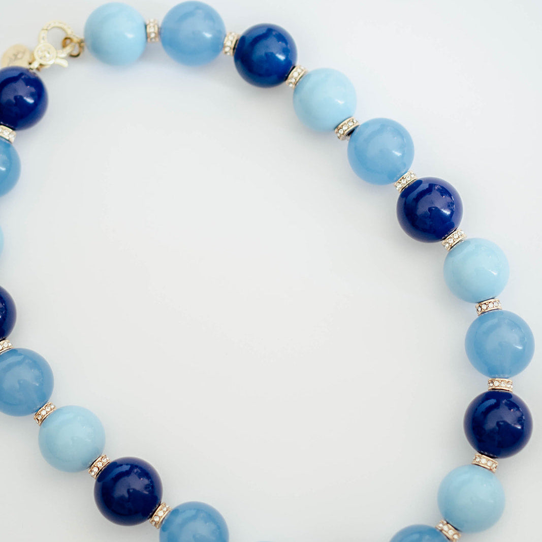 A beaded necklace made of alternating light blue and dark navy blue round beads, separated by small gold spacer beads, photographed on a clean white background.
