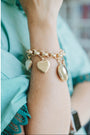 Gold bracelet with heart-shaped charms on a wrist, wearing a light blue garment.