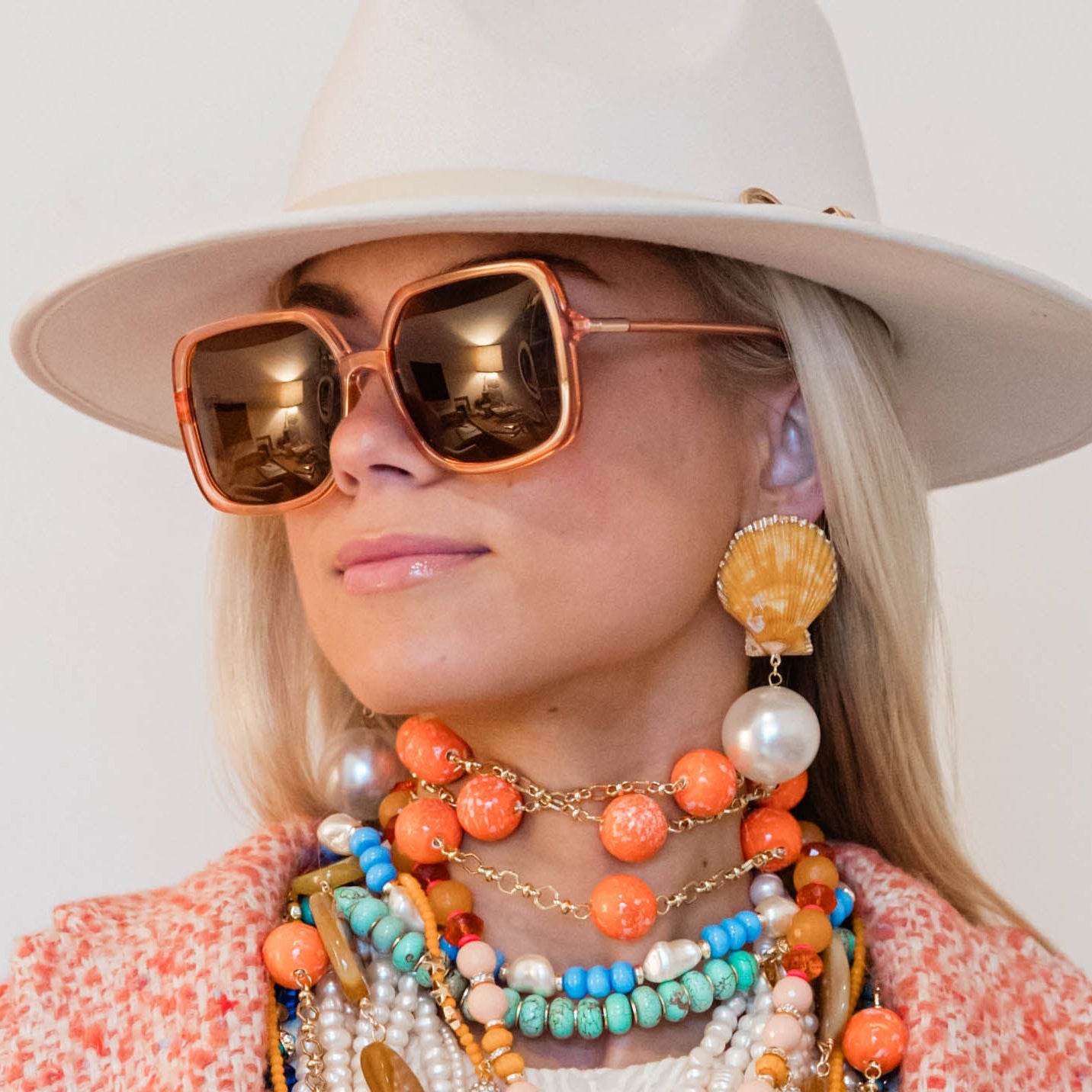Person wearing a wide-brimmed hat, sunglasses, and multiple necklaces with colorful beads against a white background