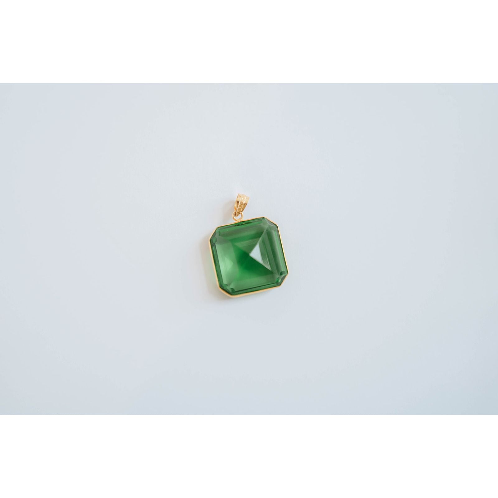 square-shaped pendant with a green crystal gem in a gold-toned brass frame against a light background