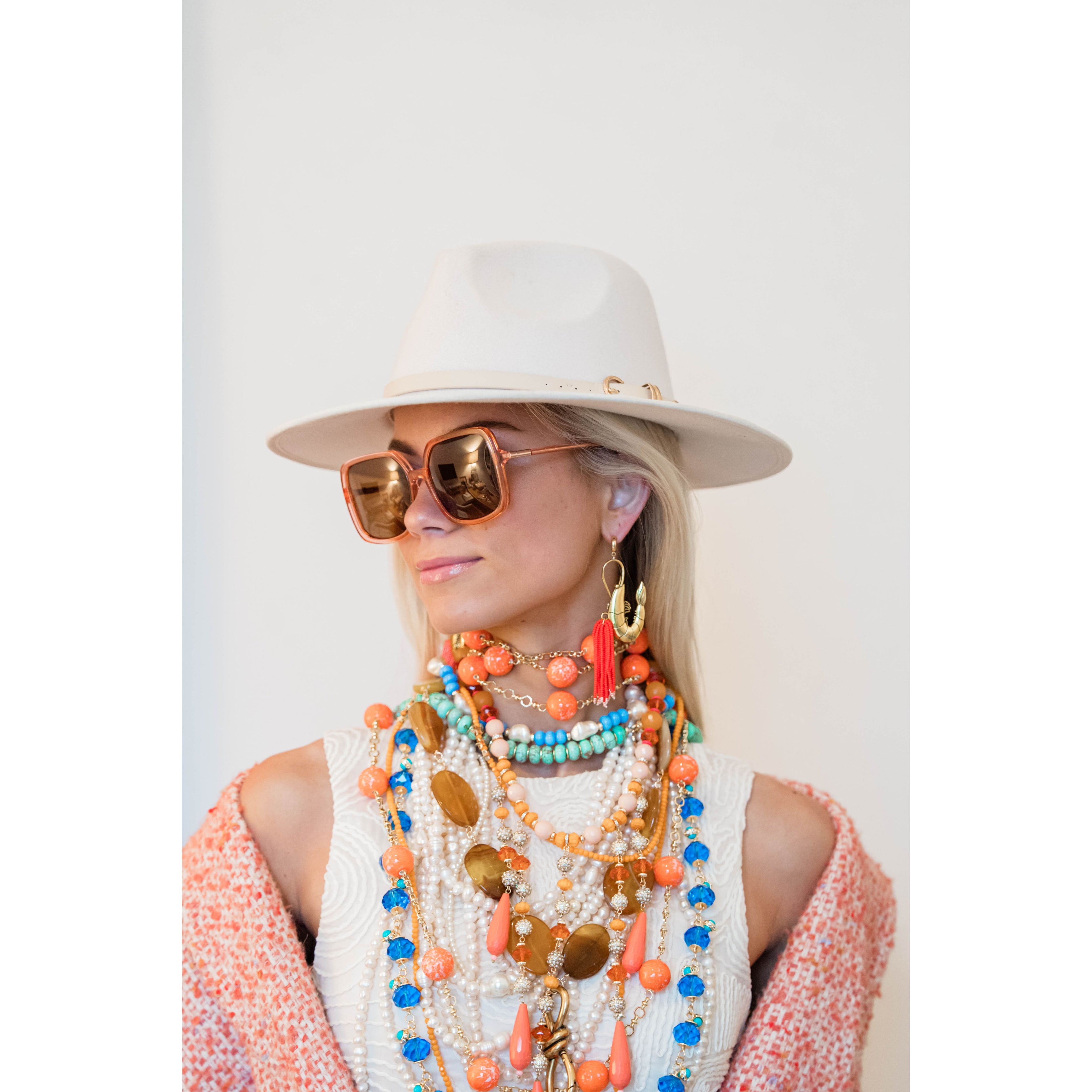 woman wearing a white hat, large sunglasses, and multiple statement beaded necklaces on a white background
