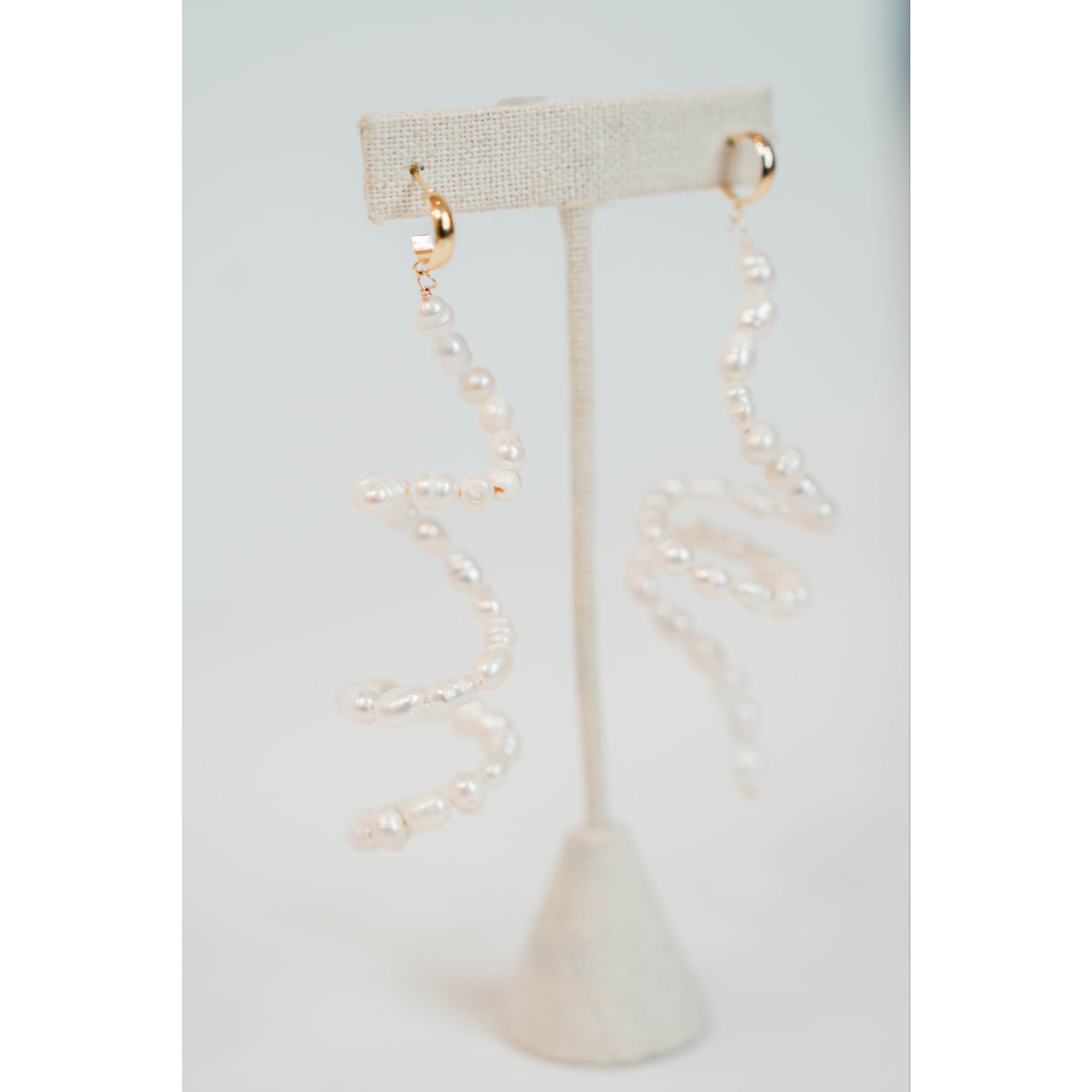 flexible pearl drop earrings with gold huggie posts on a neutral earring stand against a light gray background