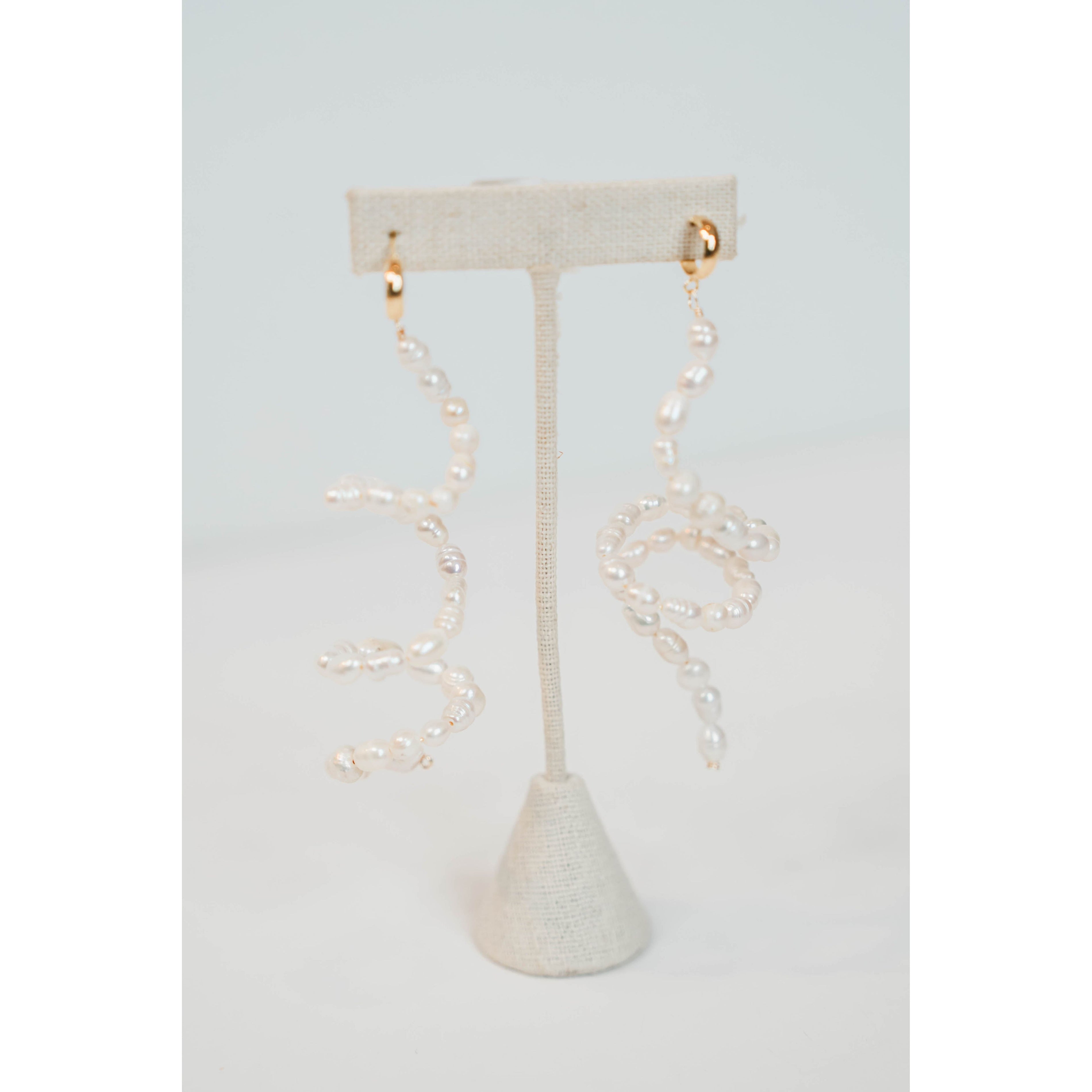 flexible pearl drop earrings with gold huggie posts on a neutral earring stand against a light gray background