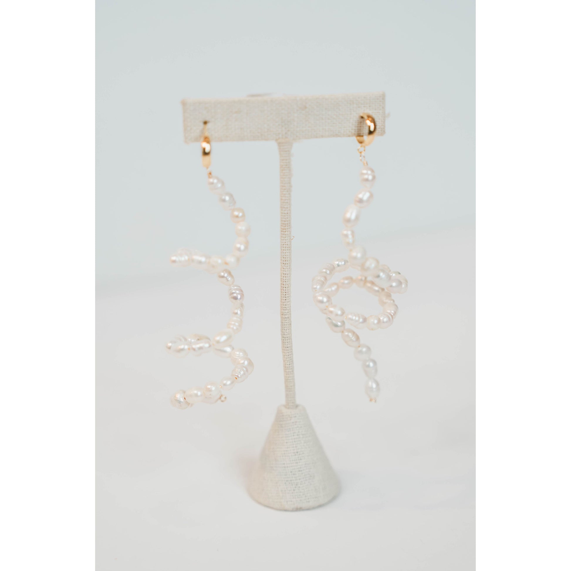 flexible pearl drop earrings with gold huggie posts on a neutral earring stand against a light gray background