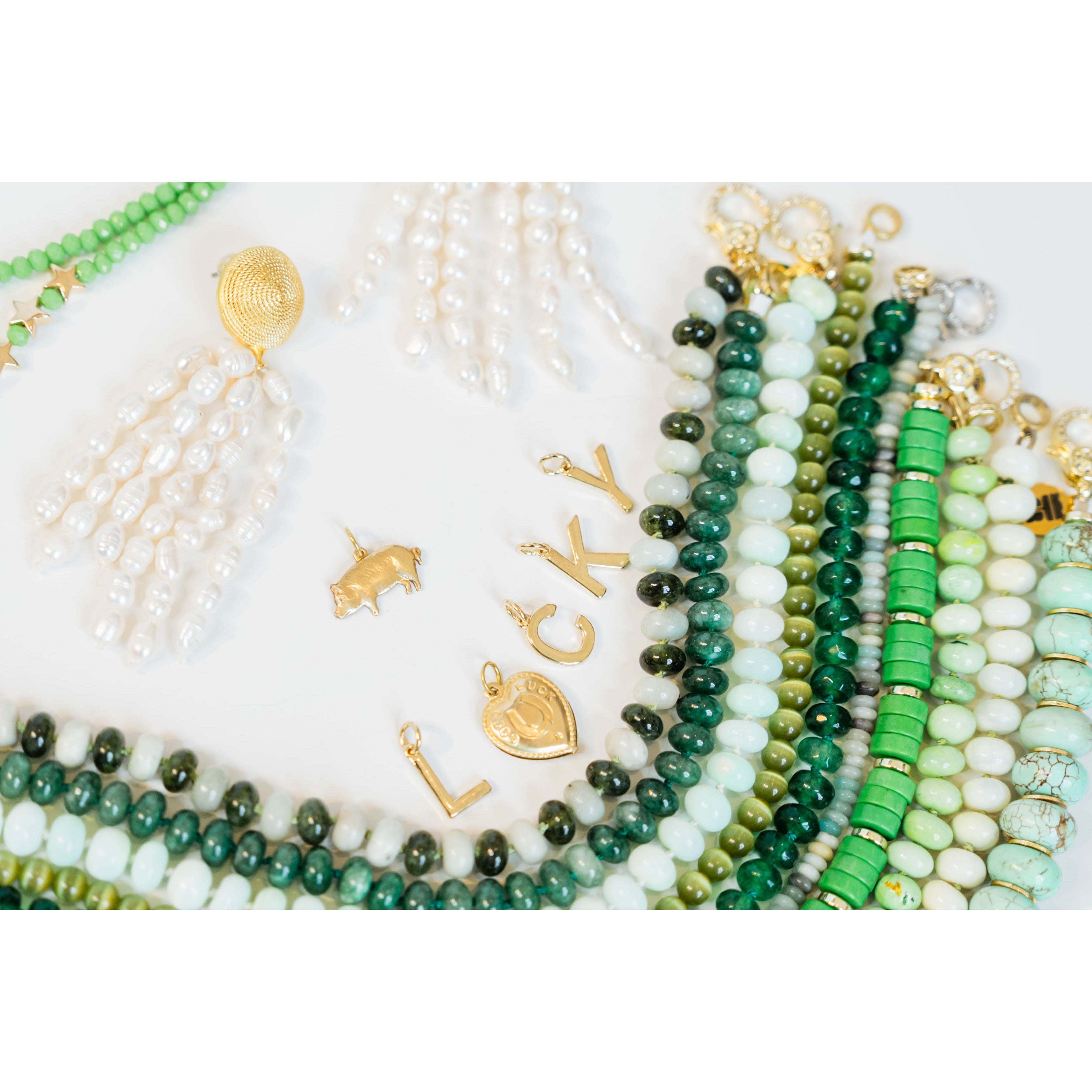 Collection of gold charms that spell out "LUCKY" and  green and white beaded necklaces and white pearl tassel earrings on a white background
