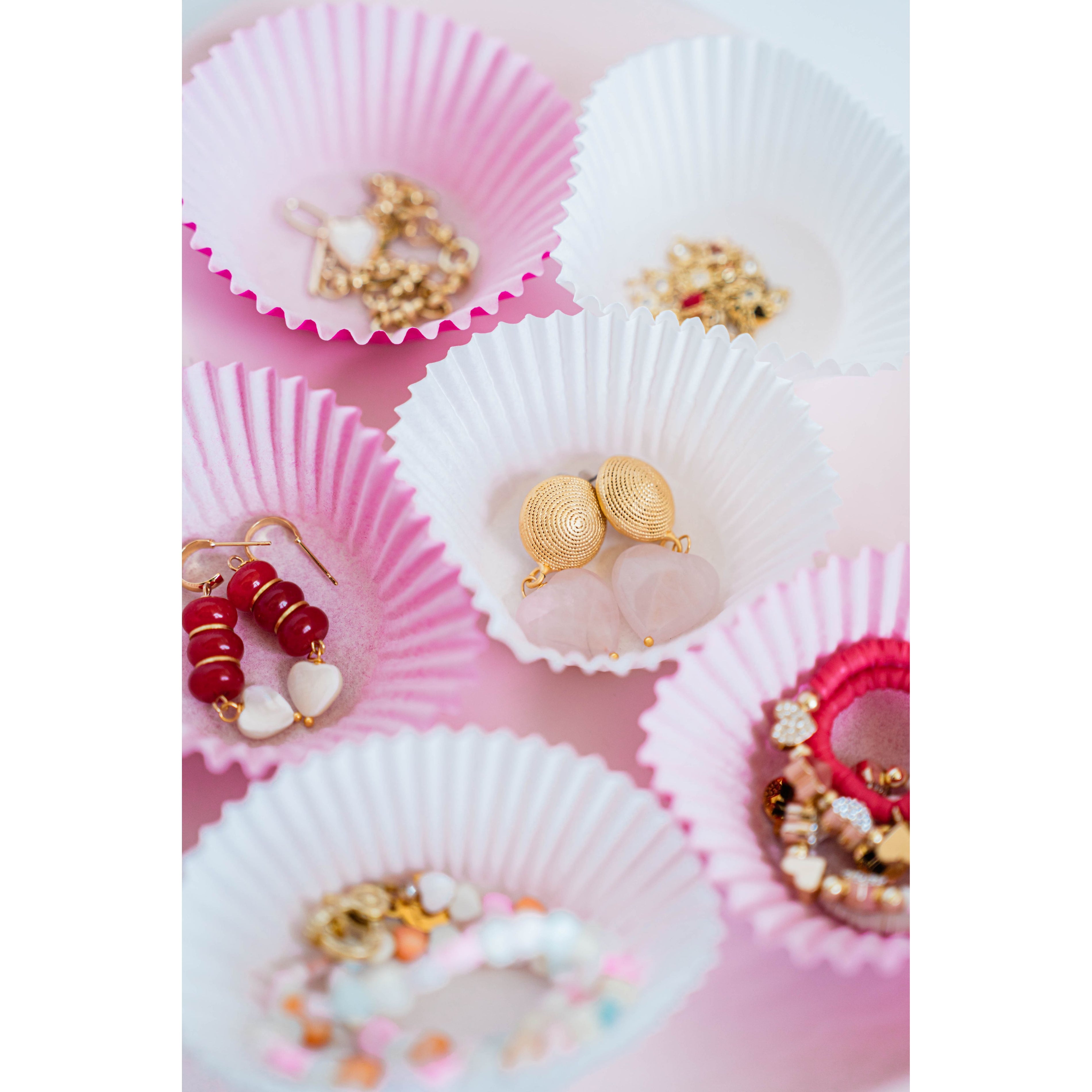 red, white, pink, and gold valentines day heart jewelry displayed in pink and white cupcake liners on a pink background